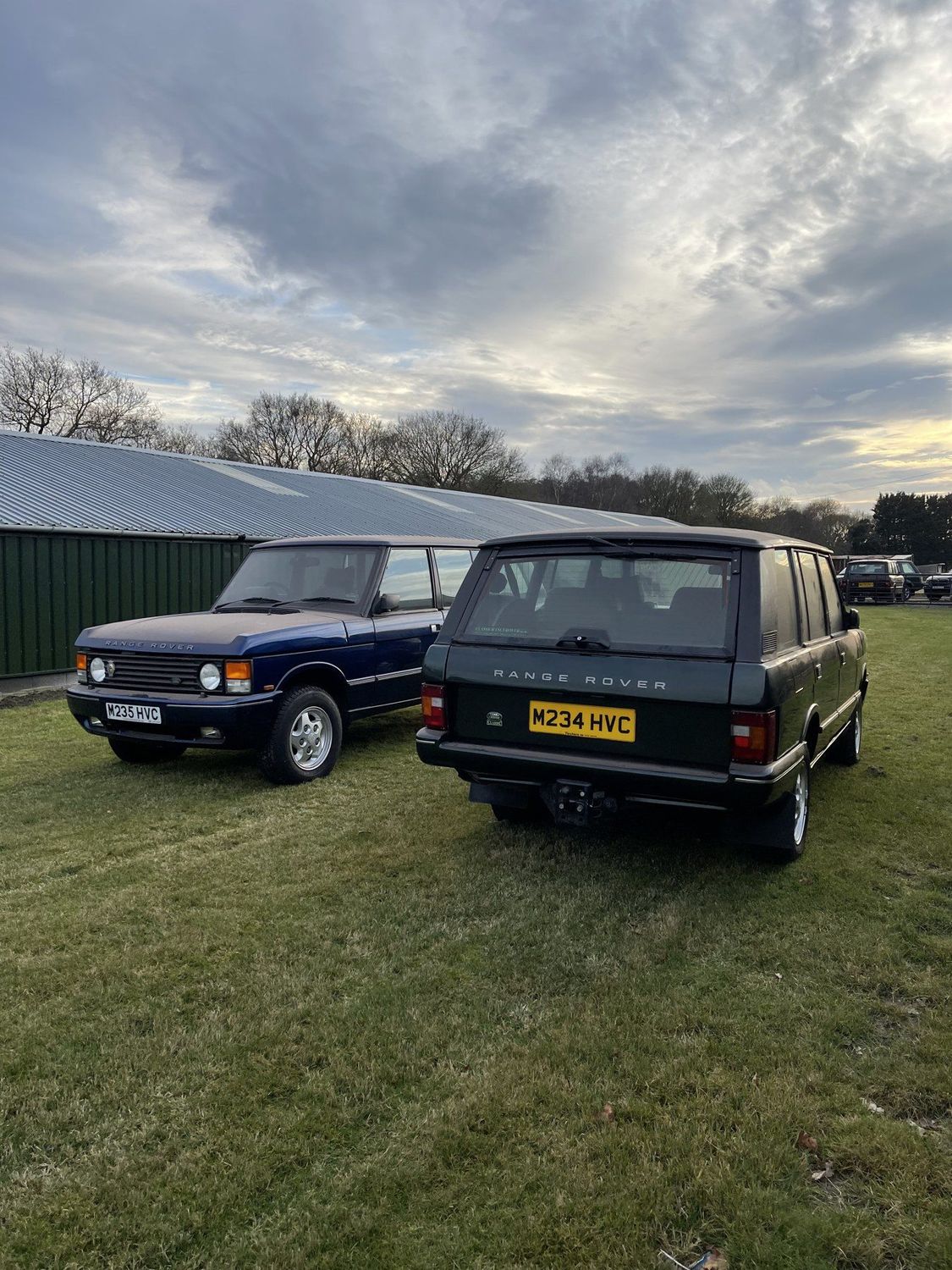 Two Range Rover Classics, one green and one navy, parked on grass at dusk